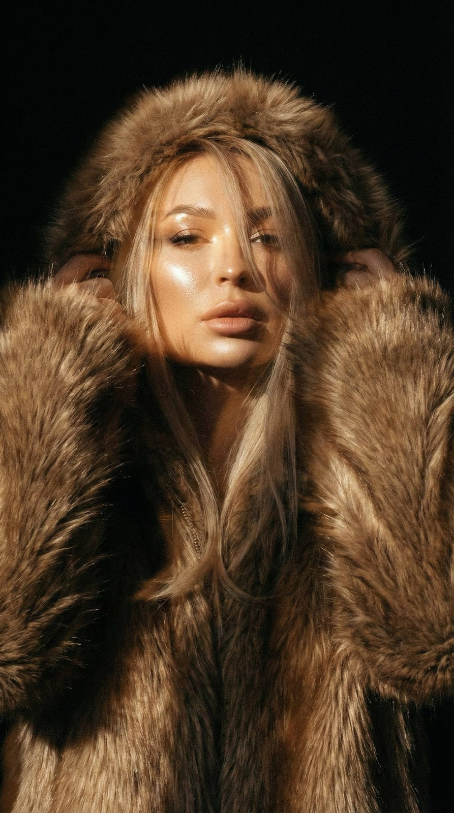 A close-up portrait of a woman in a fluffy brown fur coat, sunlight illuminating one eye, with shiny long hair partially covering her face.