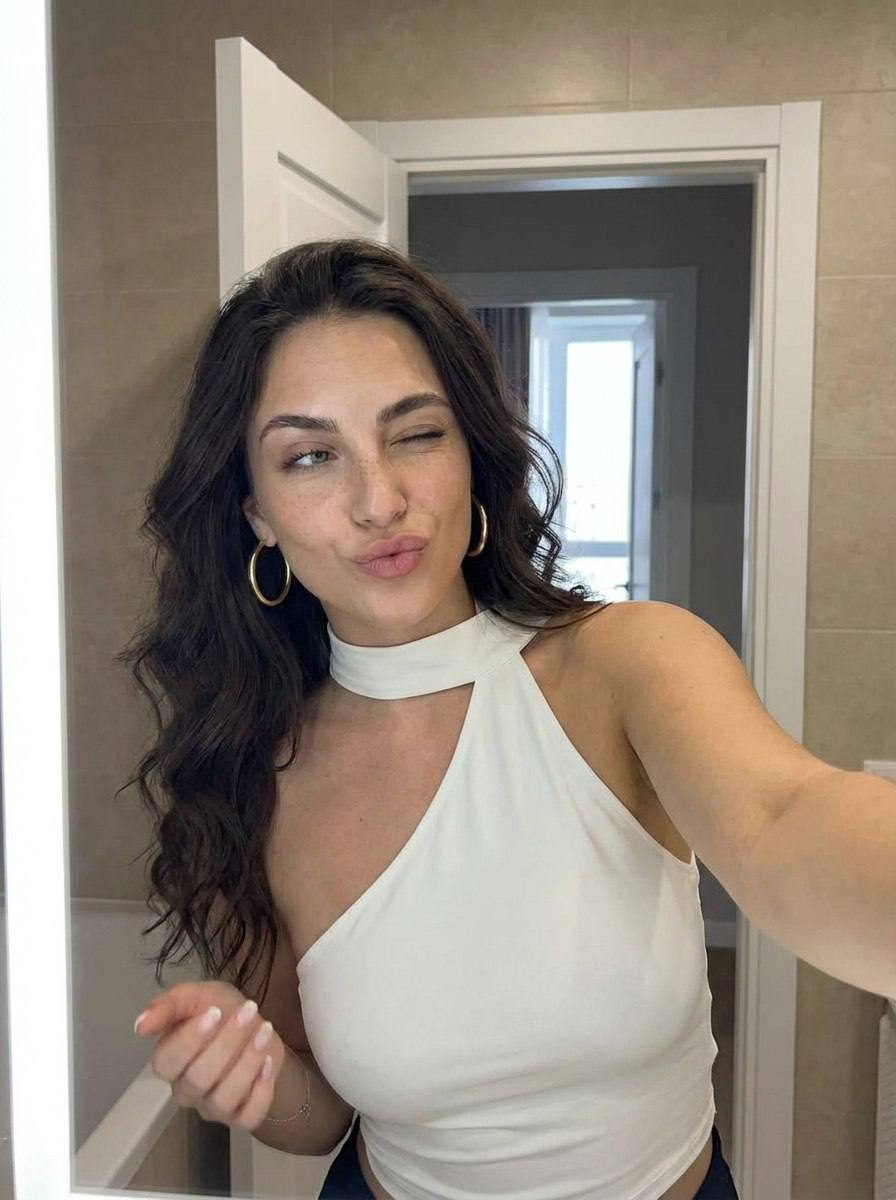 Young woman taking a selfie in a warm-lit bathroom, smiling and leaning forward, with soft curls and hoop earrings.