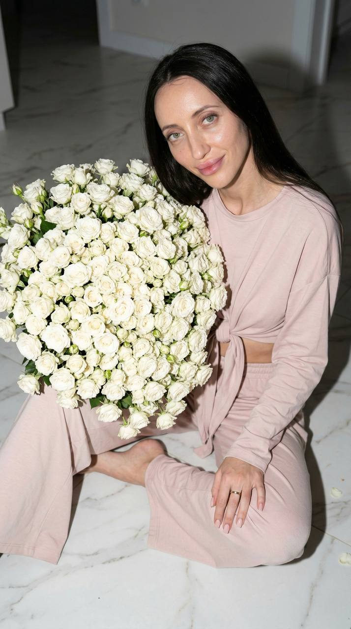 A girl in a light pink home suit sitting on a white marble floor, holding a large bouquet of spray white roses with a gentle smile.