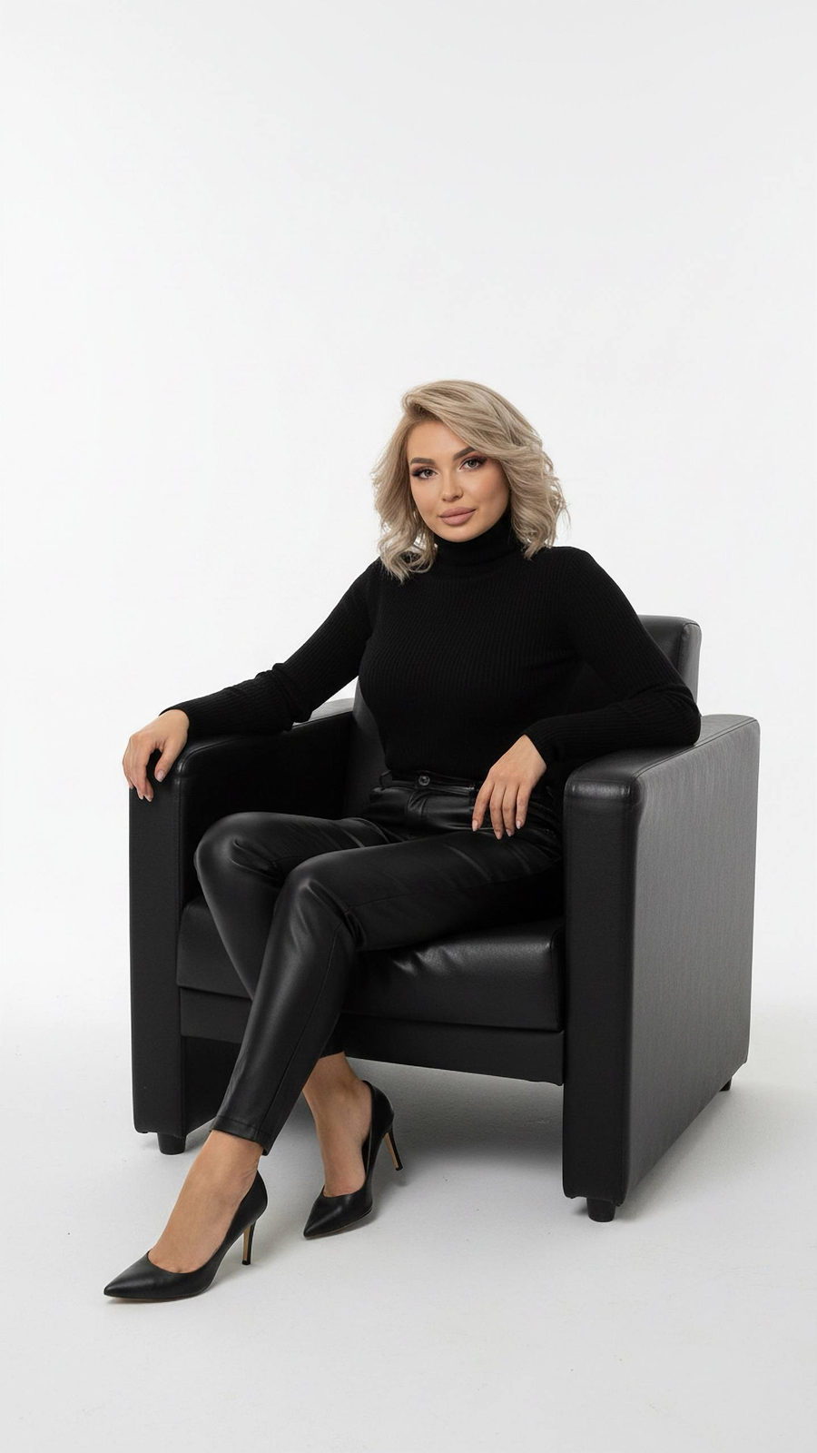 A relaxed woman in a sleek black turtleneck and leather pants sits in a modern studio, emphasizing her stylish silhouette against a minimalistic backdrop.