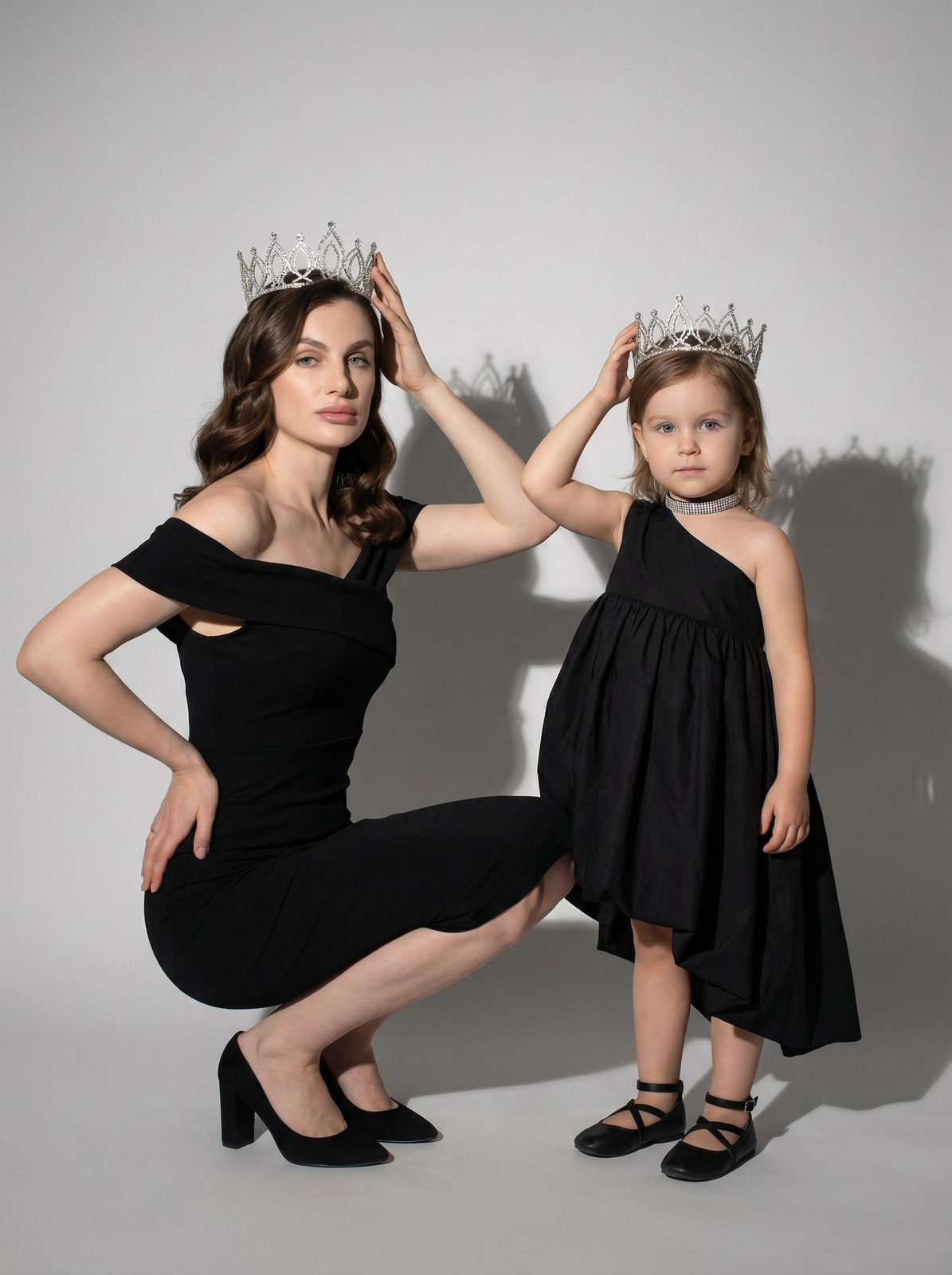 A woman in an off-the-shoulder fitted dress holding a crown while another stands beside her in a sleeveless asymmetrical dress, in a minimal studio setting.
