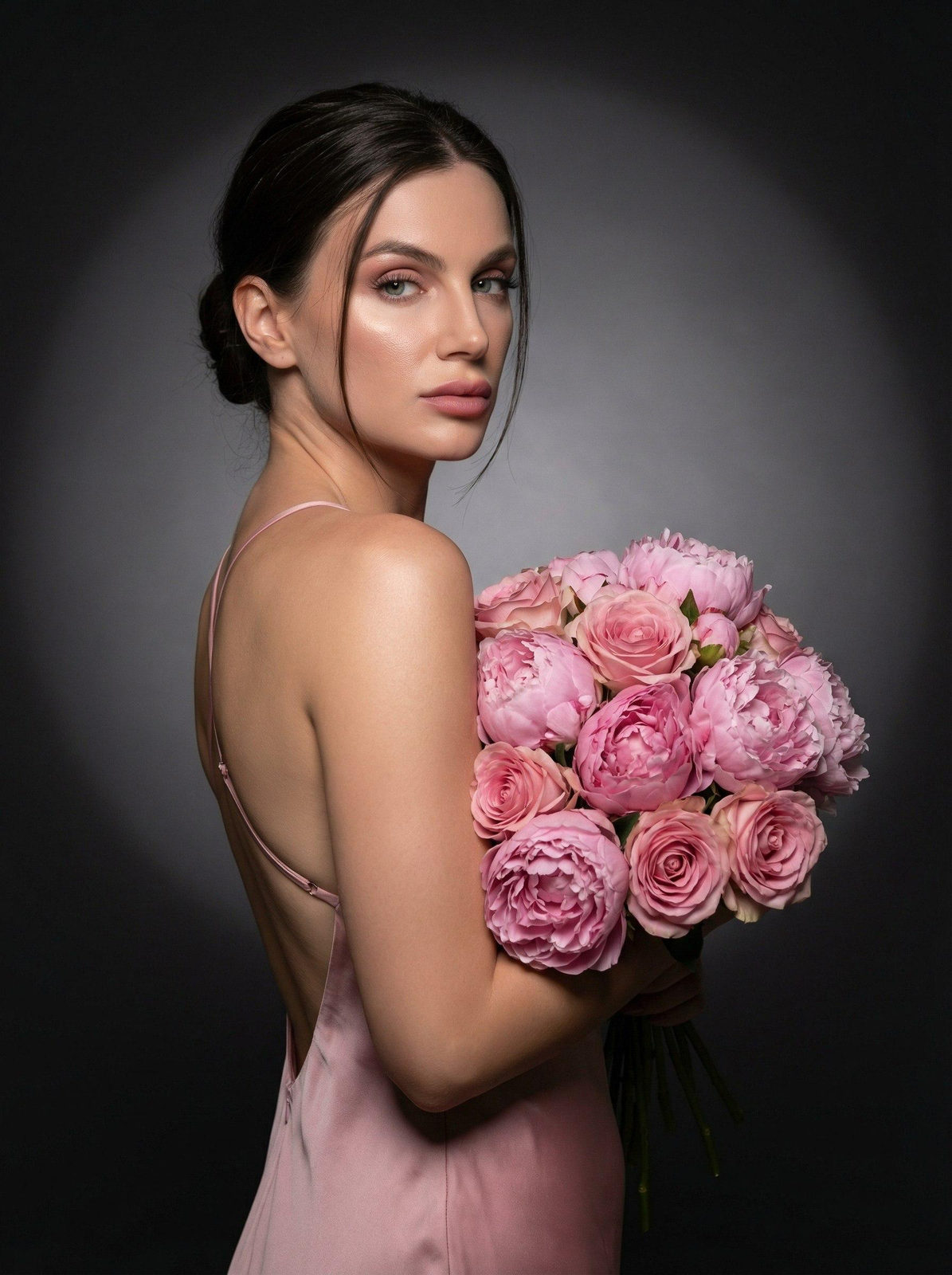 A stunning woman in a soft pink dress, holding a bouquet of pink peonies and roses in a dark studio setting.
