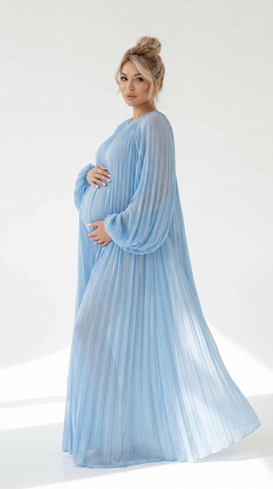 Pregnant woman standing gracefully in a light blue dress against a large white backdrop, with soft lighting and flowing fabric.