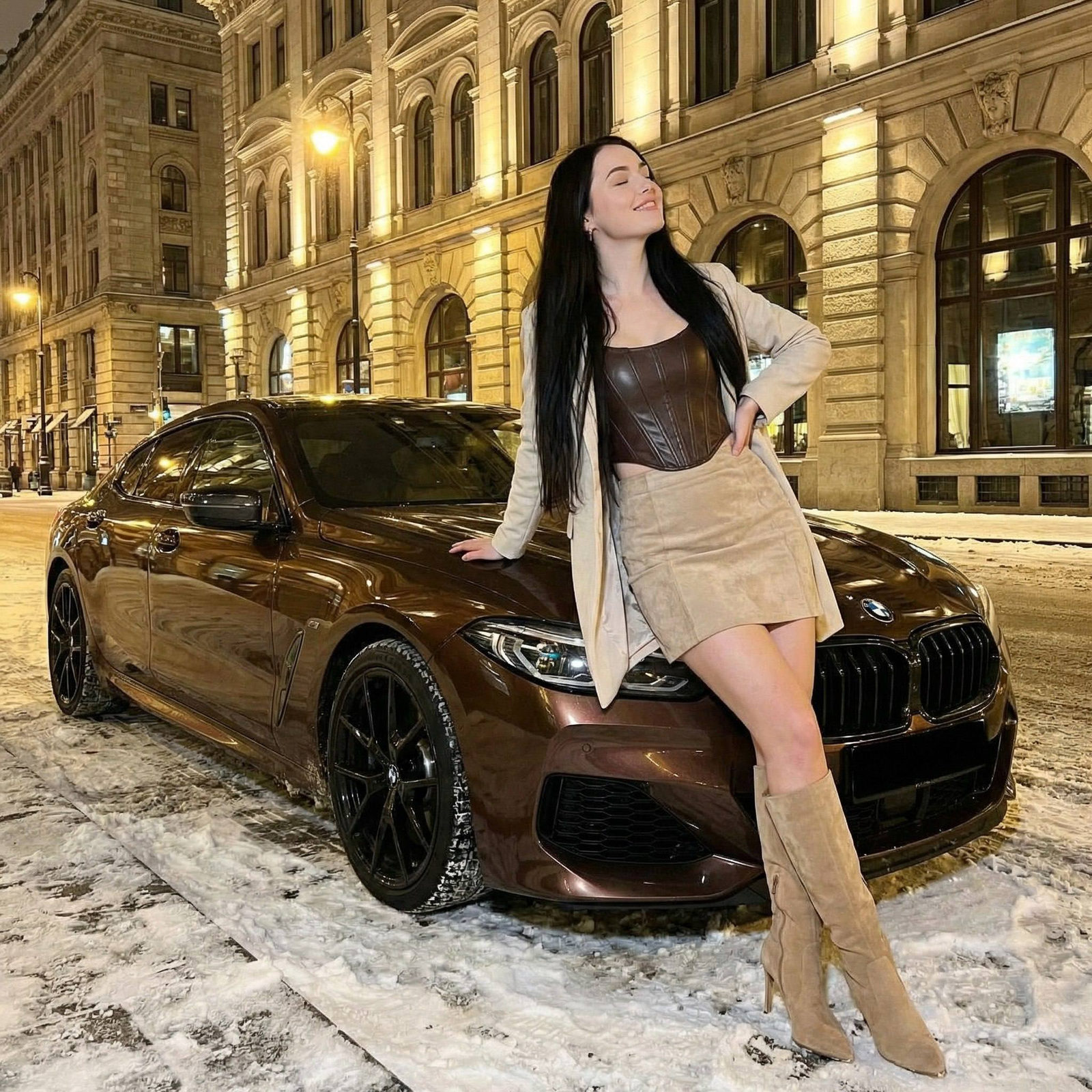 A stylish young woman in a dark brown corset top and beige skirt poses confidently by a luxury BMW on a snowy city street at night.