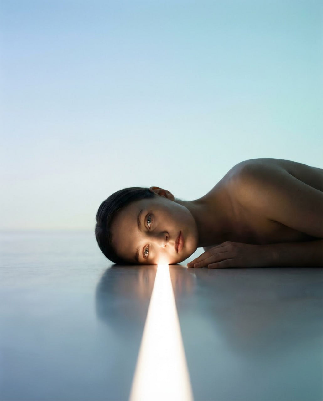 A surreal minimalist portrait of a person lying on a smooth floor, illuminated by a bright stripe of light highlighting their facial features.