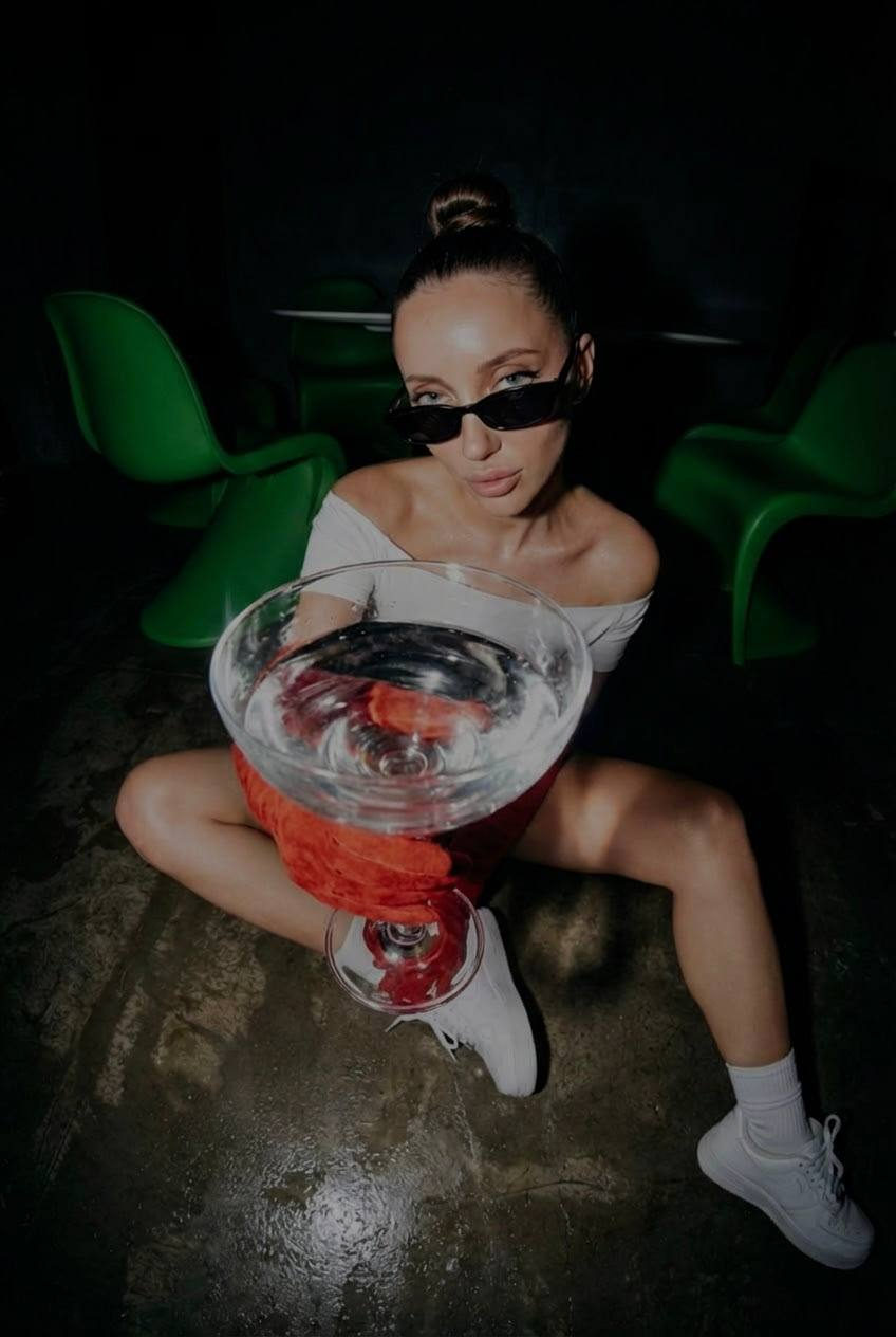 A girl sitting on a shiny concrete floor in a dark club, holding a large transparent cocktail glass, wearing narrow y2k sunglasses.