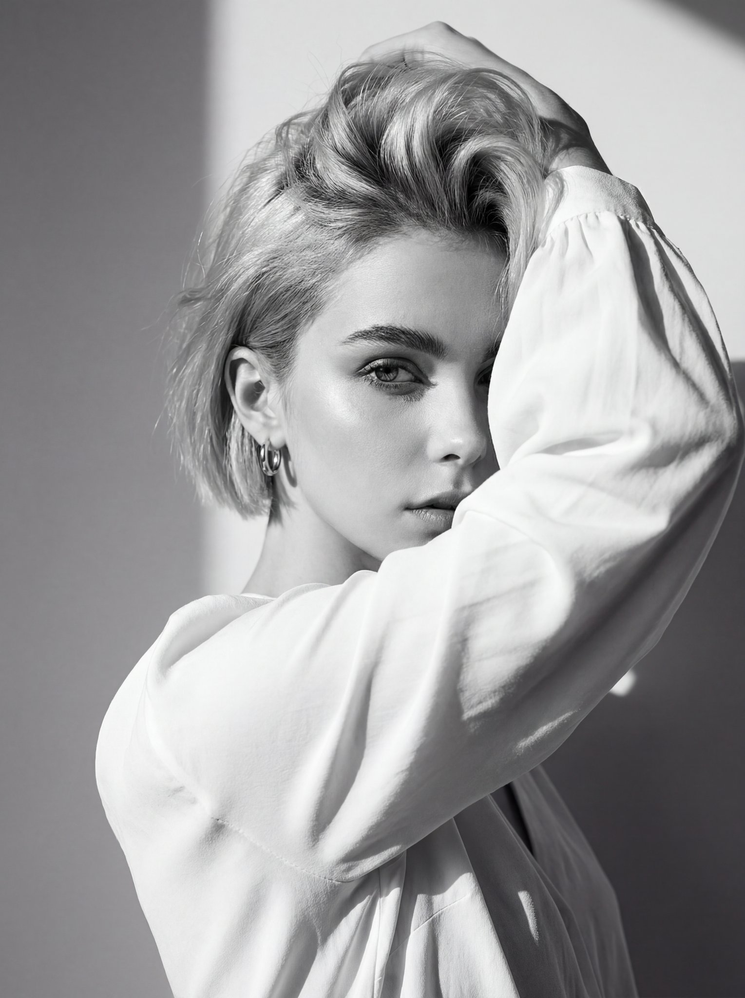 Black and white editorial portrait of a woman in a white blouse, her arm raised partially covering her face, showcasing sharp shadows and dramatic lighting.