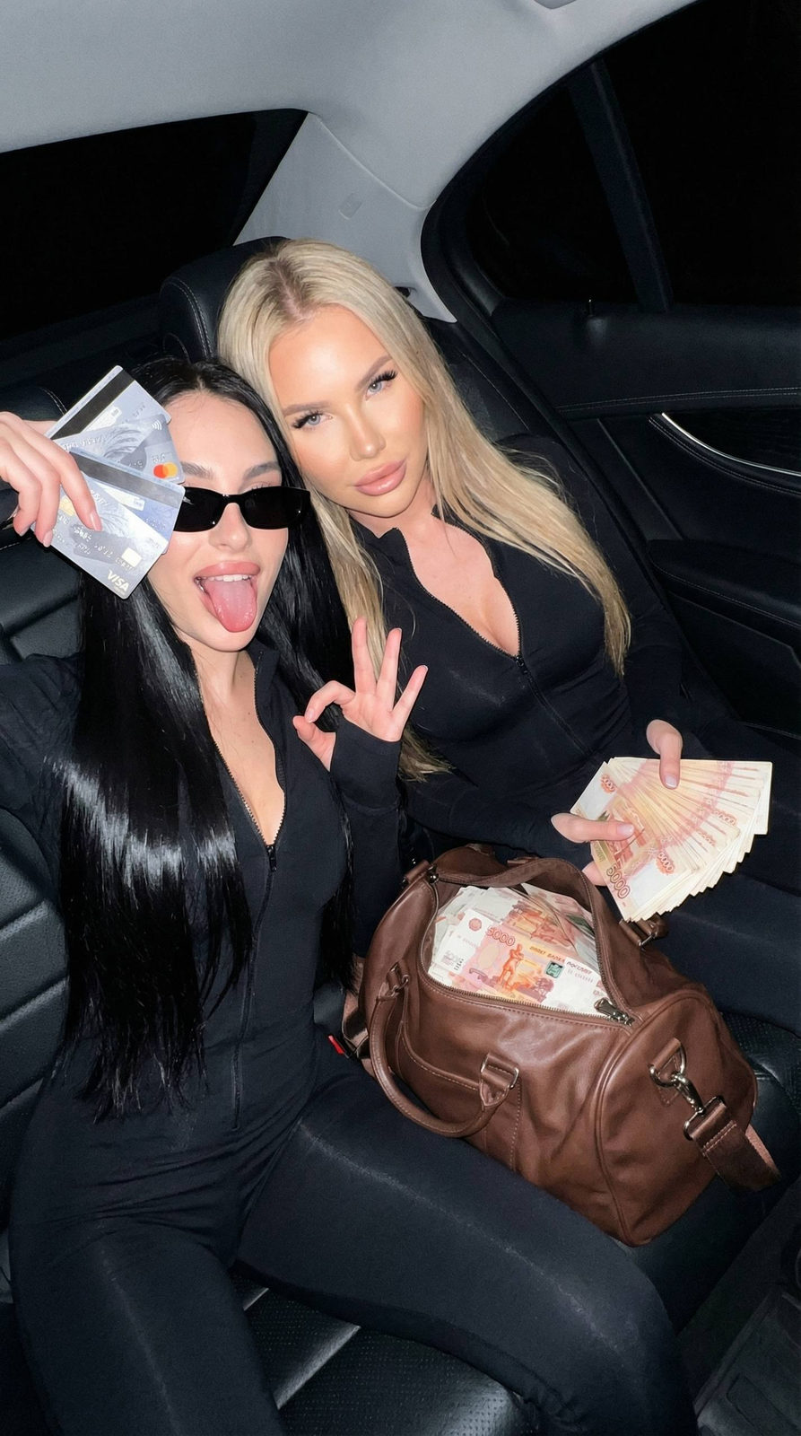 A stunning scene inside a luxury car with two girls in black jumpsuits, one holding money while the other poses playfully.