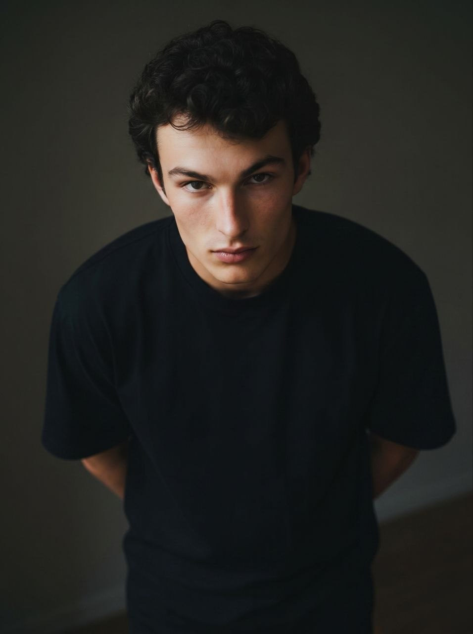 A man in a dimly lit room, leaning forward with a serious expression, wearing a loose black t-shirt, sharp jawline and soft shadows.