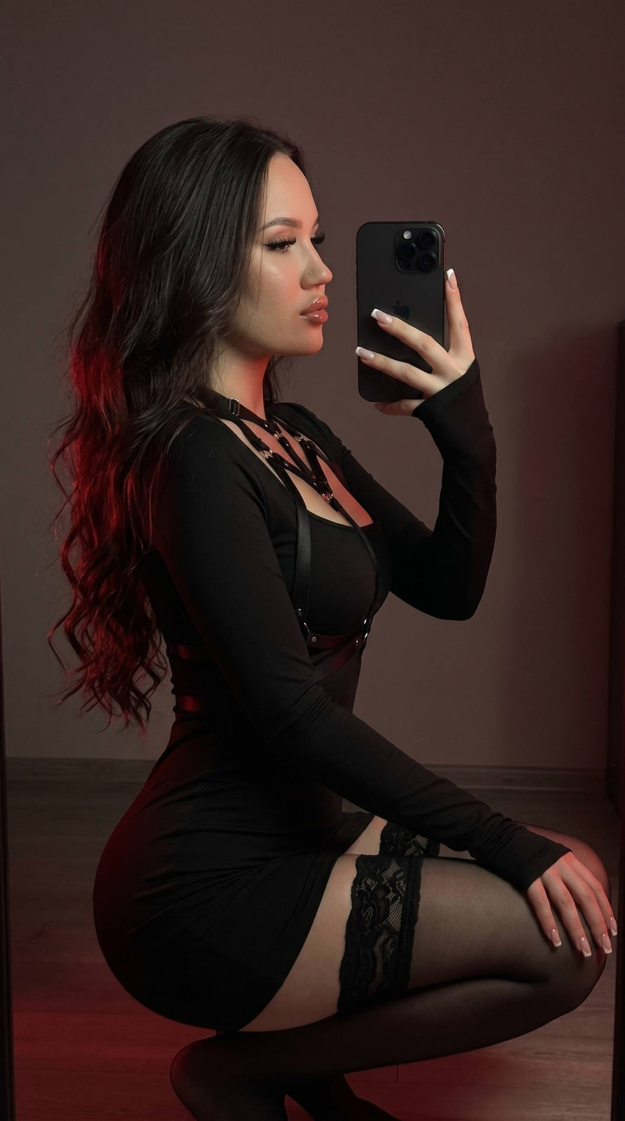 A woman with long glossy wavy hair, wearing a fitted black dress, hand partially covering her face while posing in dim lighting.