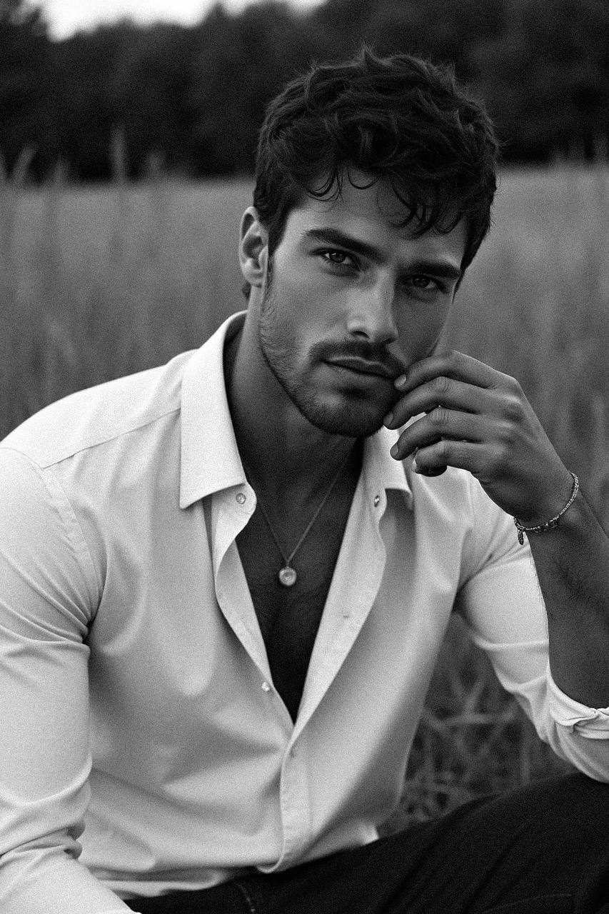 A young man sitting outdoors in a field, gazing at the camera with a calm expression, wearing a slightly unbuttoned shirt, captured in monochrome.