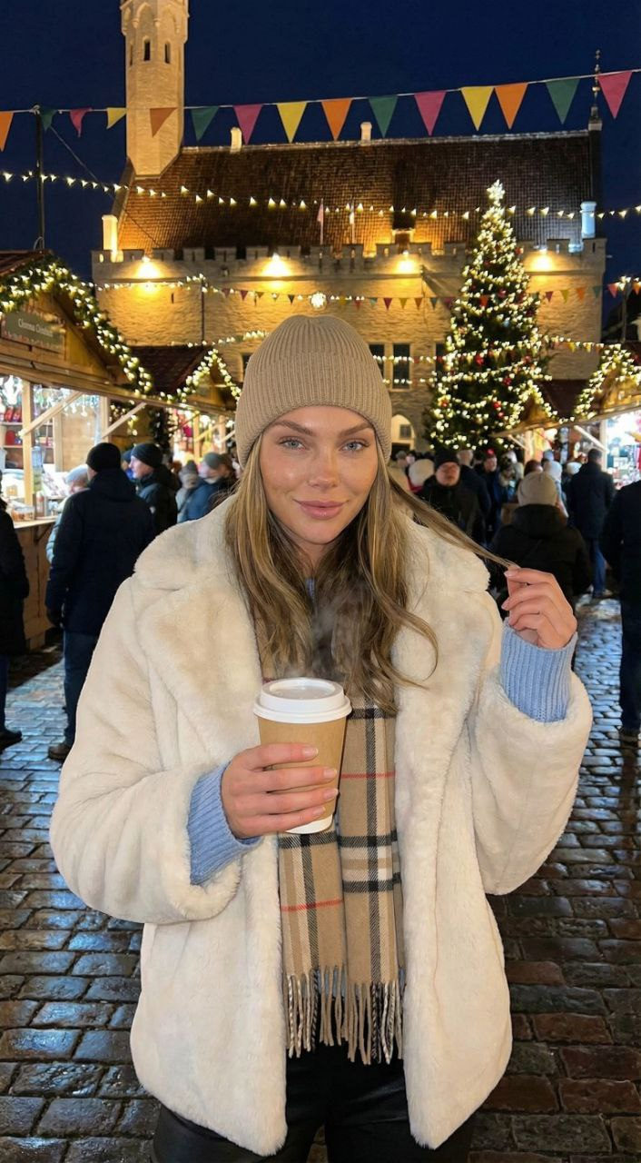 A woman in a cozy faux-fur coat stands in a vibrant Christmas market, holding a hot drink, surrounded by holiday lights and decorations.
