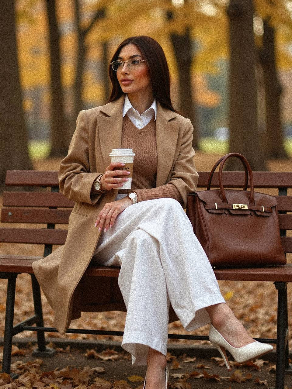 Ultra-detailed autumn lifestyle portrait of a woman in a chic outfit, sitting on a park bench surrounded by falling golden leaves.