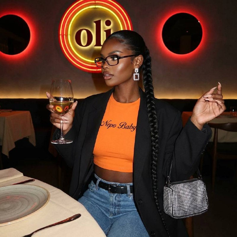 A stylish woman in an orange cropped top and dark jacket, posing in a modern restaurant with a glass of wine and sparkling accessories.