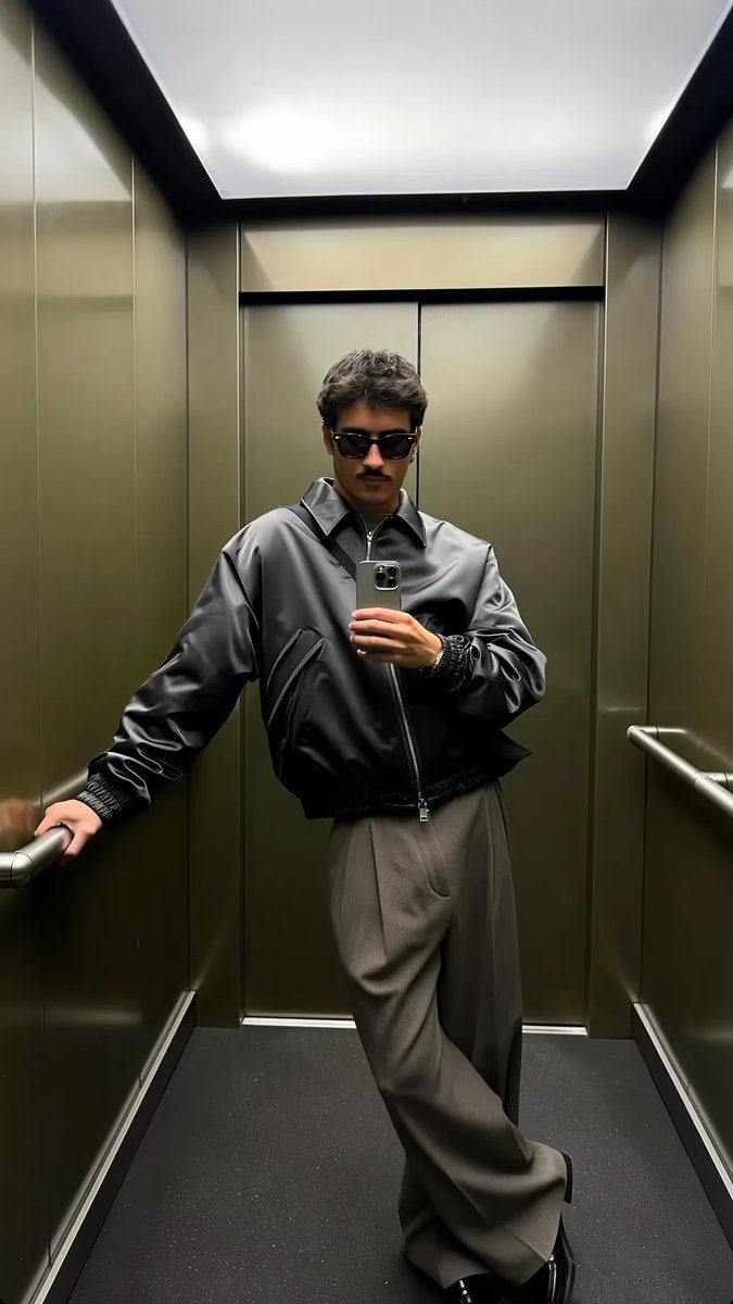 A stylish man in an olive shadowed elevator, arms stretched, reflecting on the glass with cool rails in the backdrop.