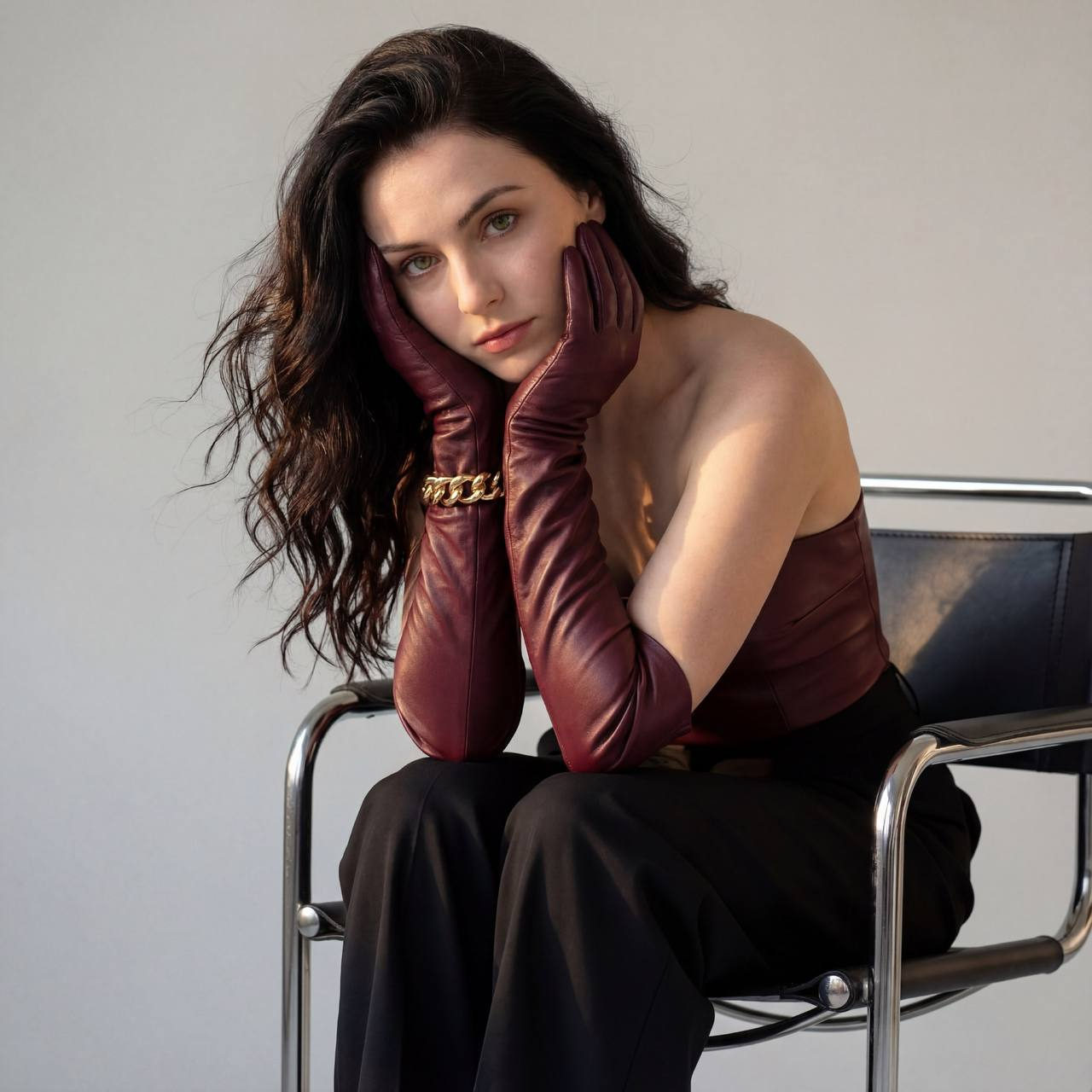 A woman in a burgundy leather top and gloves, sitting on a modern metal chair, with hair flowing and soft lighting highlighting her thoughtful expression.
