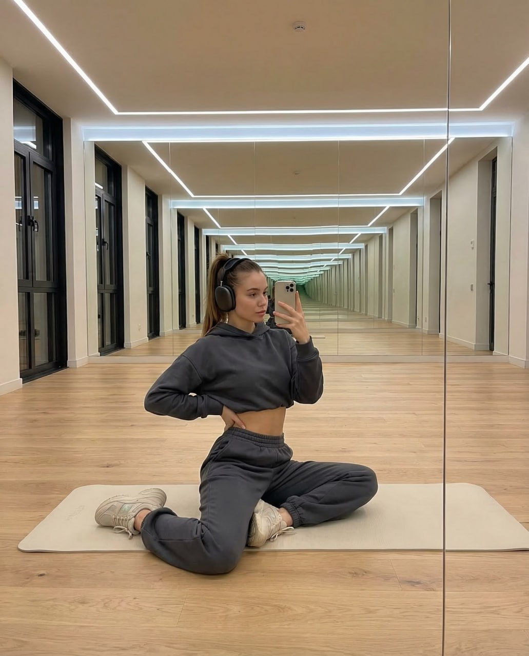 A girl in a cropped graphite hoodie poses in front of a mirror, showcasing a relaxing Pilates studio setting.