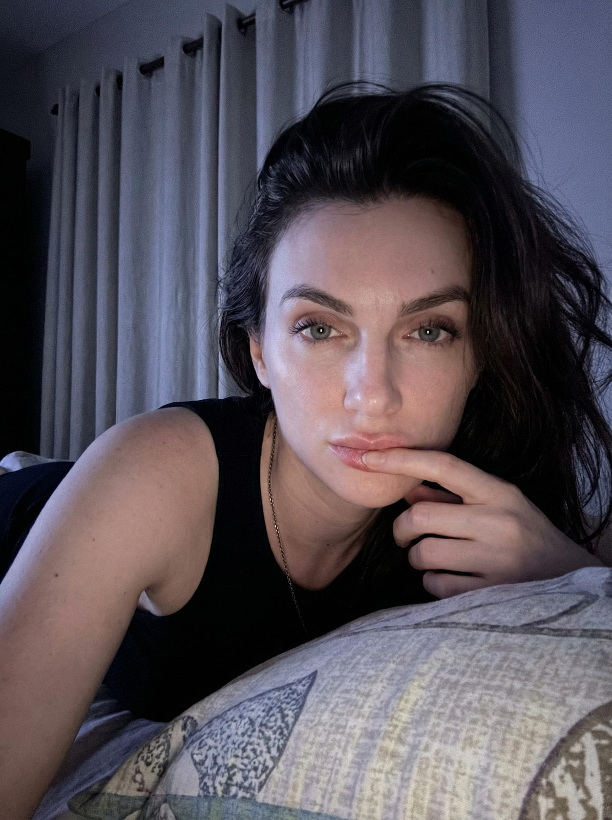 Close-up of a woman lying in bed, her left hand touching her lip, with soft, cool lighting highlighting her facial features.