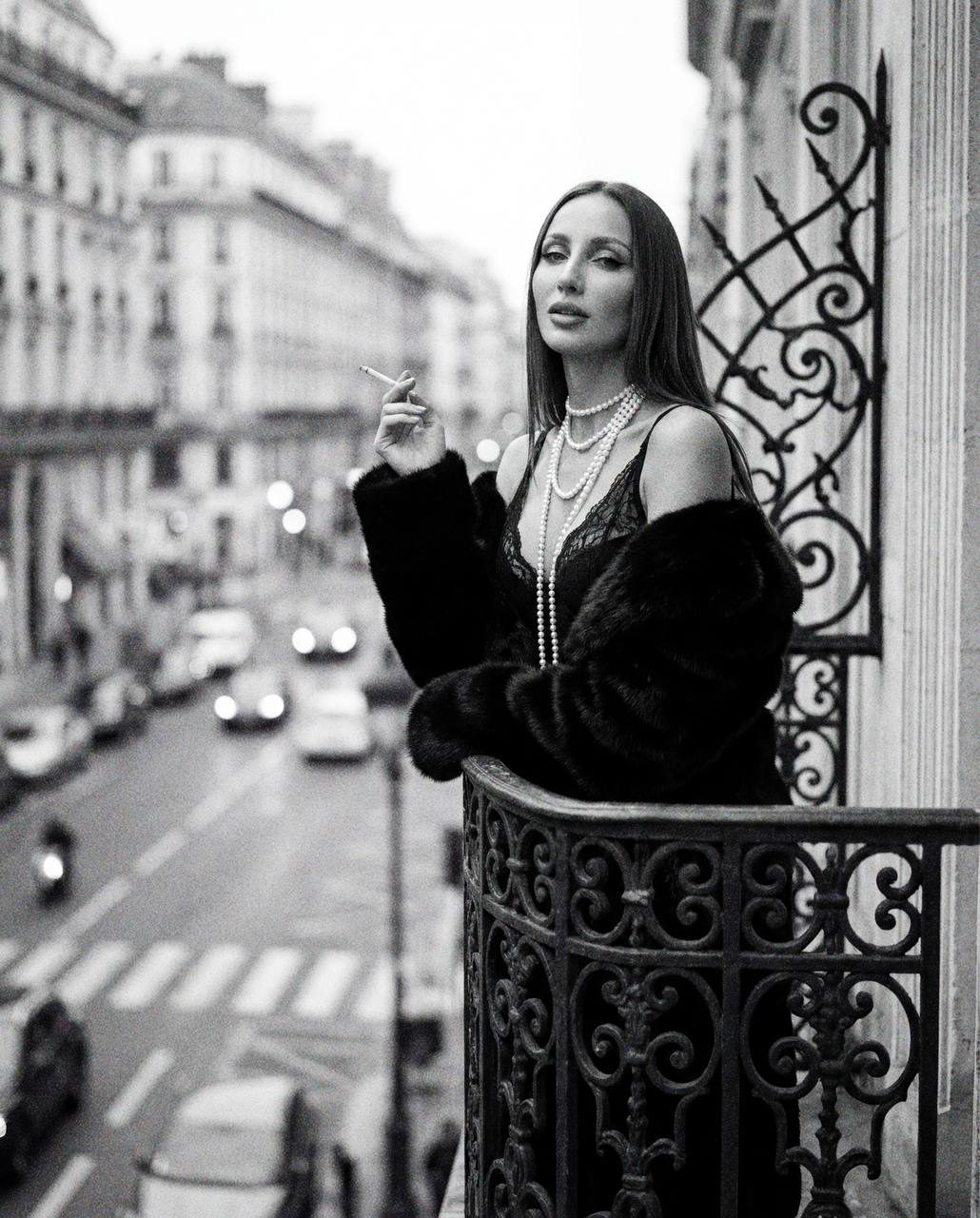 A stylish woman in a lace top and fur coat elegantly poses on a balcony with a cigarette, showcasing vintage Parisian charm.
