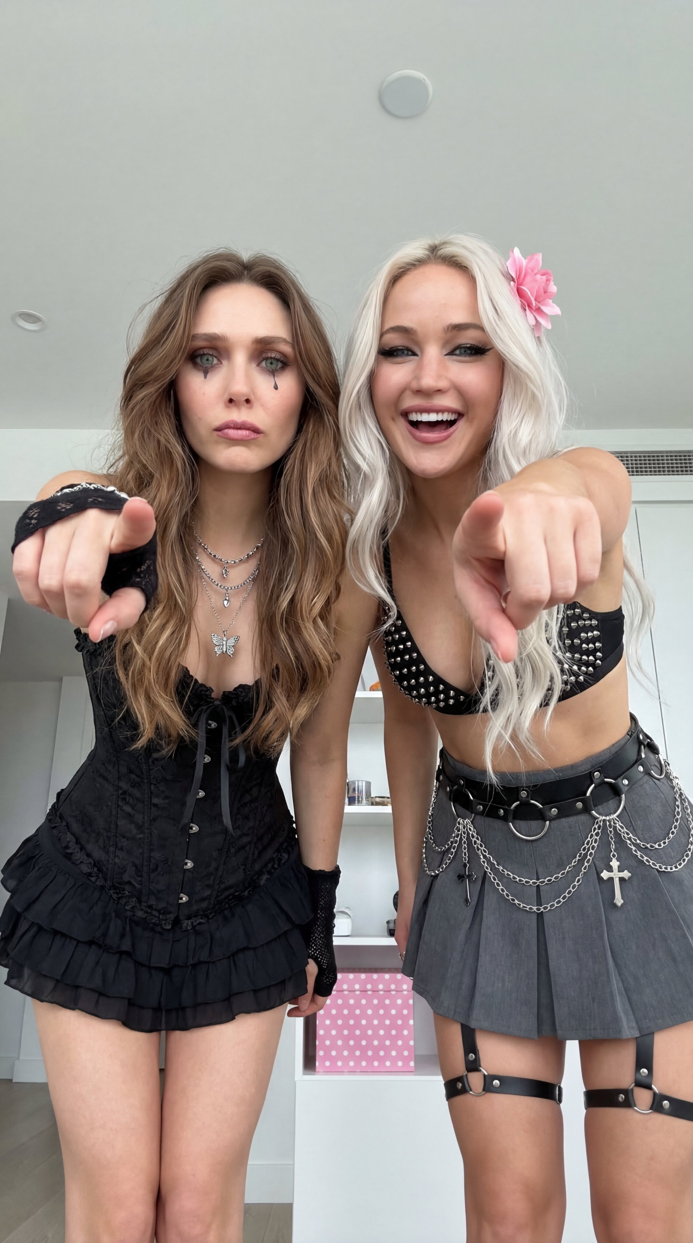 Two individuals in gothic outfits pointing at the camera, set in a bright room.
