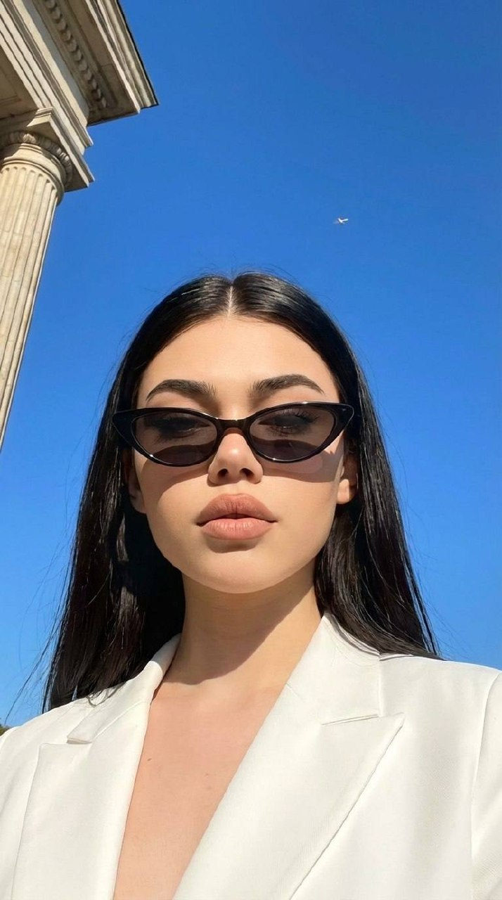 A beautiful young woman in a white blazer and stylish sunglasses against a bright blue sky, confident expression and sunny lighting.