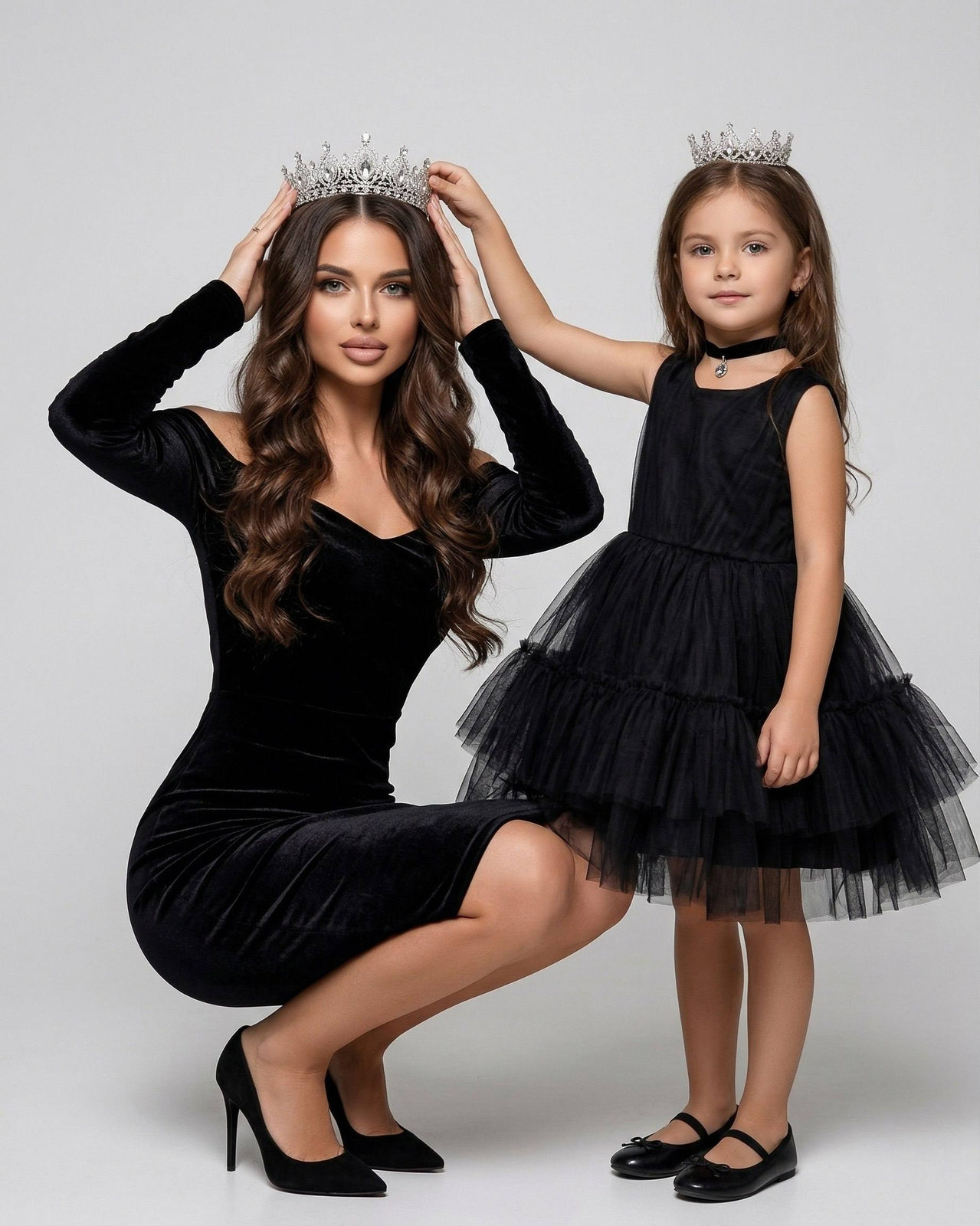 A woman in an off-shoulder black dress squats on the left holding a silver crown, while a child in a full-skirt black dress stands on the right, both looking at the camera.