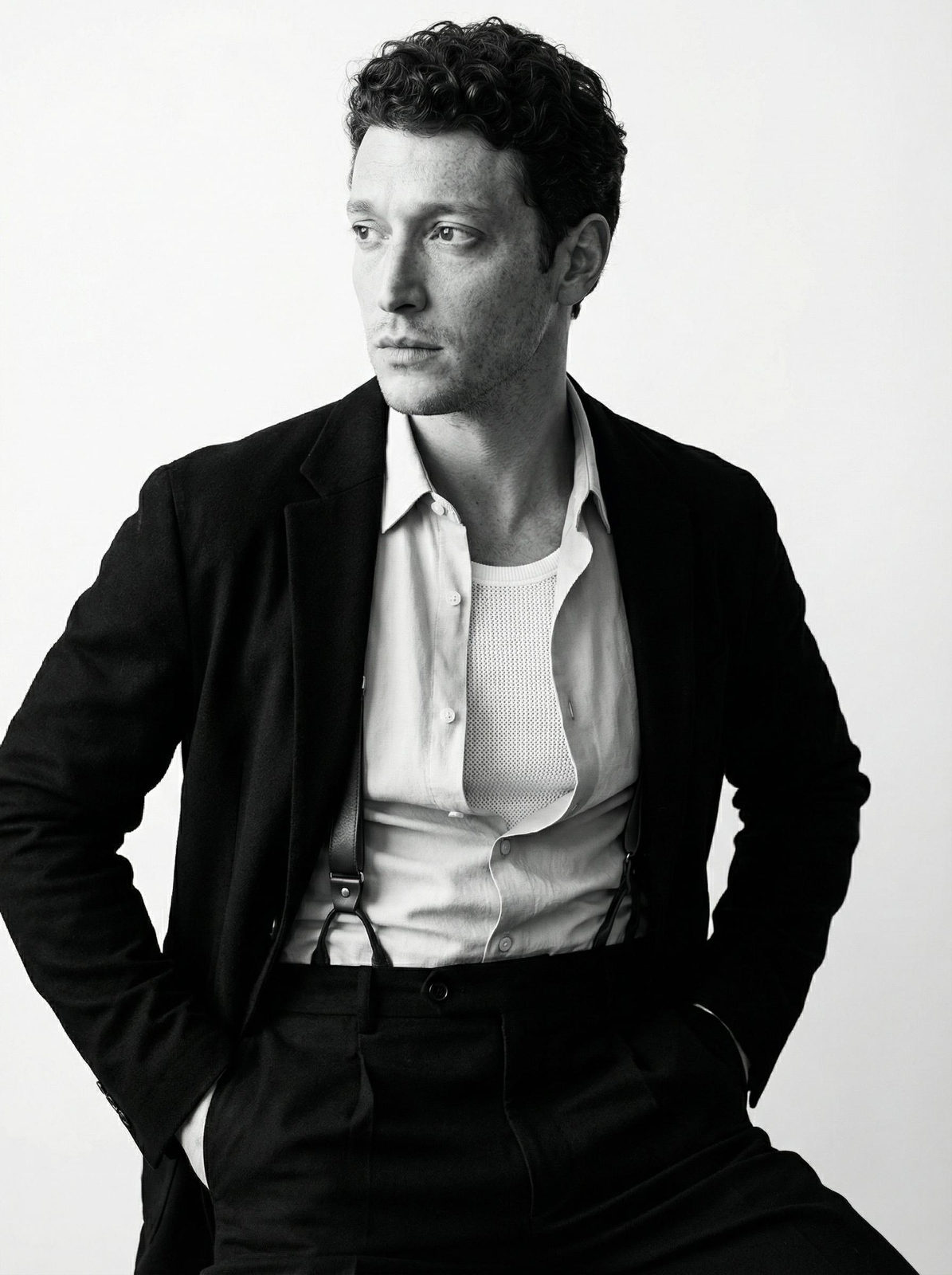 A confident man in a classic black jacket and light shirt stands with hands in pockets against a minimalist white background.