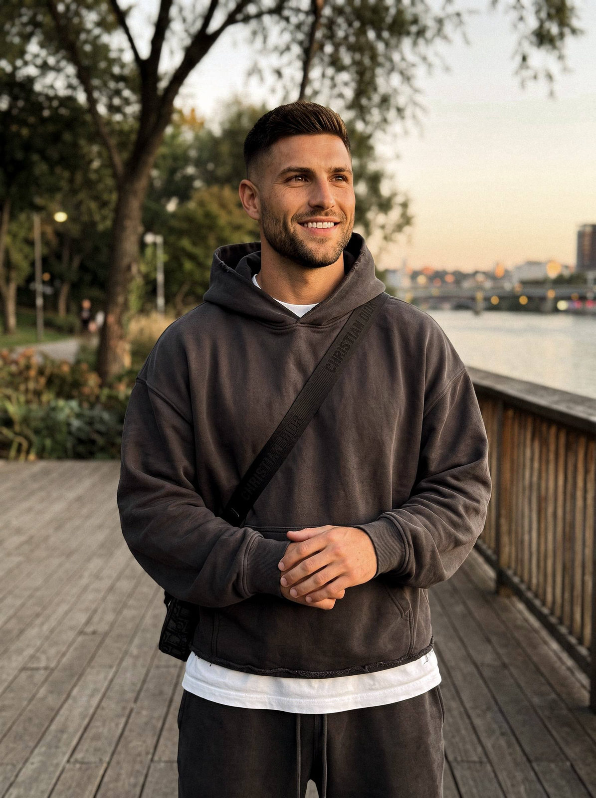 A man in a dark hooded sweatshirt and crossbody bag smiling softly in a park setting.